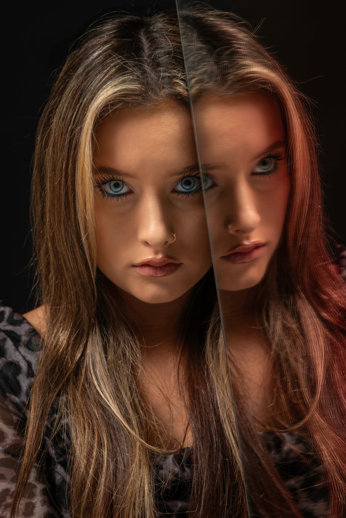 woman with blue eyes looking strait on with her reflection in a mirror she is next to. studio photography.