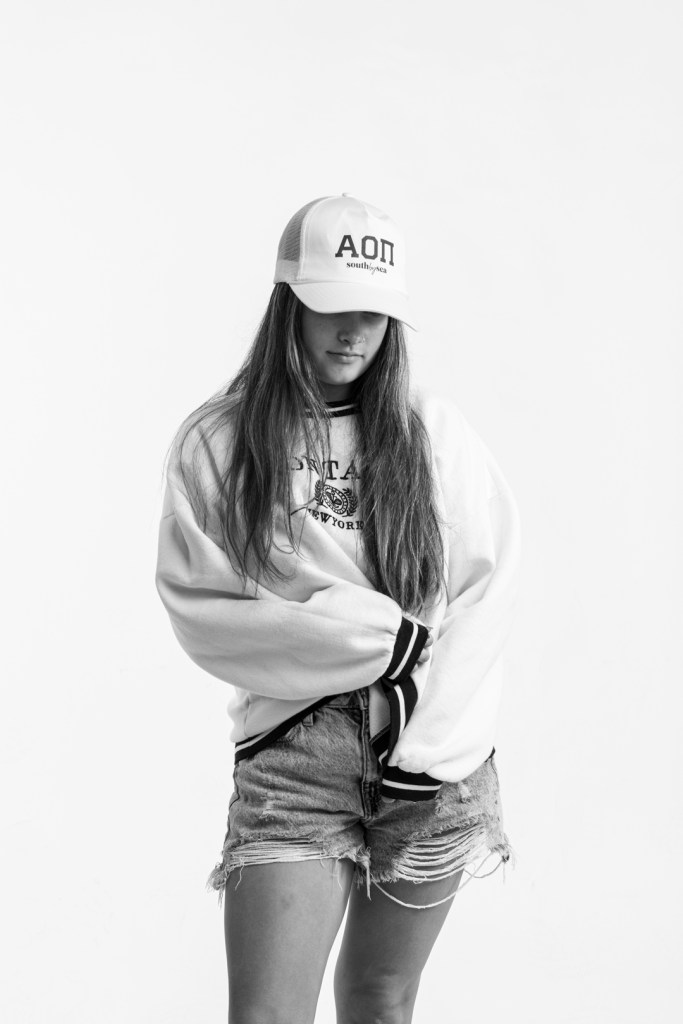 woman in white hat and sweatshirt looks down and tugs at the bottom of her sweatshirt. studio photography.