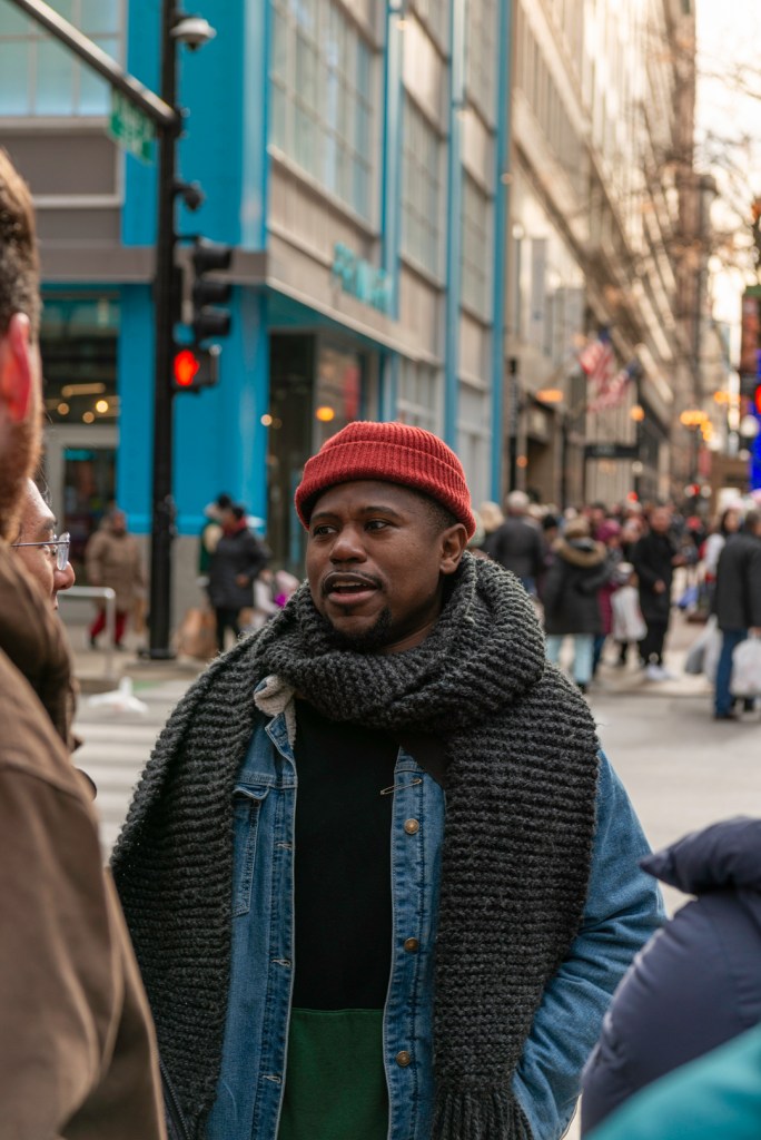 Man stands on the sidewalk with a red has and a large scarf.